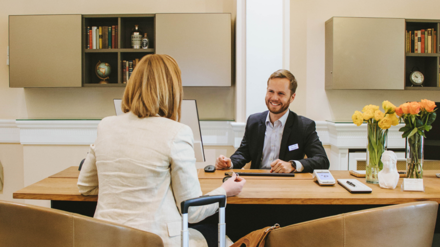 Check-In an der Rezeption im Louisa´s Place Hotel. Schick gekleidete Frau sitzt mit Koffer an einem Tisch und wird vmo Rezeptionisten freundlich empfangen.