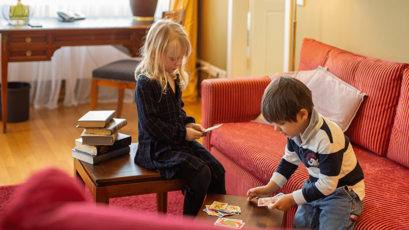 Zwei Kinder, ein Junge und ein Mädchen sitzen im Wohnzimmer der Louisa´s Suite im Louisas Place Hotel in Berlin. Die Kinder spielen Karten. Das Sofa ist rot.