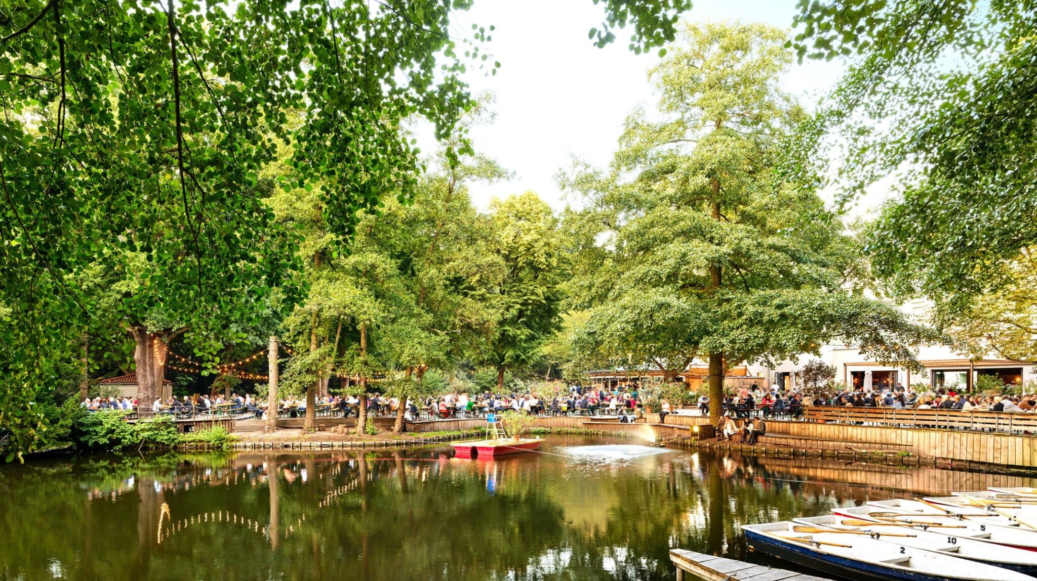 Aufnahme vom Café am Neuen See im Tiergarten in Berlin. Vorne liegen Boote im Wasser, mittig ein kleiner See, drumherum Bäume und im Hintergrund Menschen auf Bierbänken.